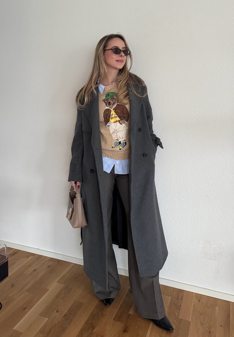 Manteau long gris porté sur un pull beige avec un motif d'ours, associé à une chemise bleu clair, un pantalon marron, des chaussures noires à bout pointu, et un petit sac.
