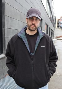 Homme portant une veste Carhartt noire avec col en flanelle à carreaux et casquette Oakley grise, debout sur un trottoir urbain près de murs de bâtiment gris.
