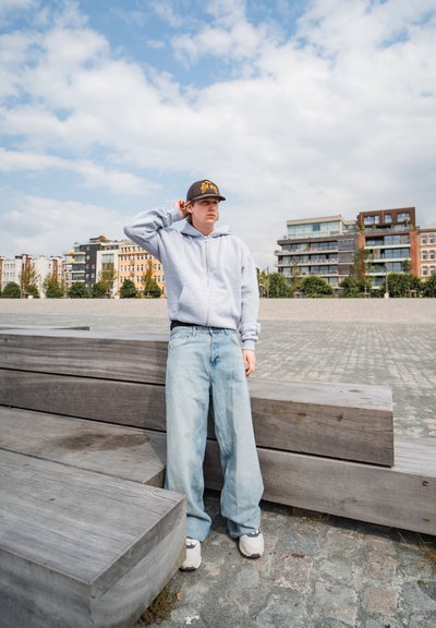 Sudadera gris con cremallera, jeans holgados azul claro y zapatillas blancas. De pie junto a bancos de madera, con una gorra negra que tiene un logo amarillo.
