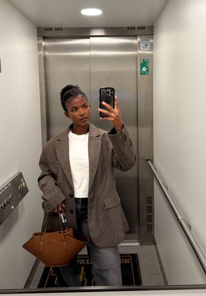 Blazer marrón de gran tamaño sobre una camiseta blanca, combinado con jeans de cintura alta. Un bolso estructurado marrón con asas, dentro de un ascensor.