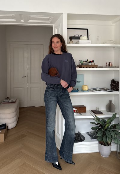 Mujer de pie en interiores con suéter oscuro, vaqueros acampanados y zapatos puntiagudos, sosteniendo un bolso pequeño marrón, con estanterías blancas y decoración detrás de ella.