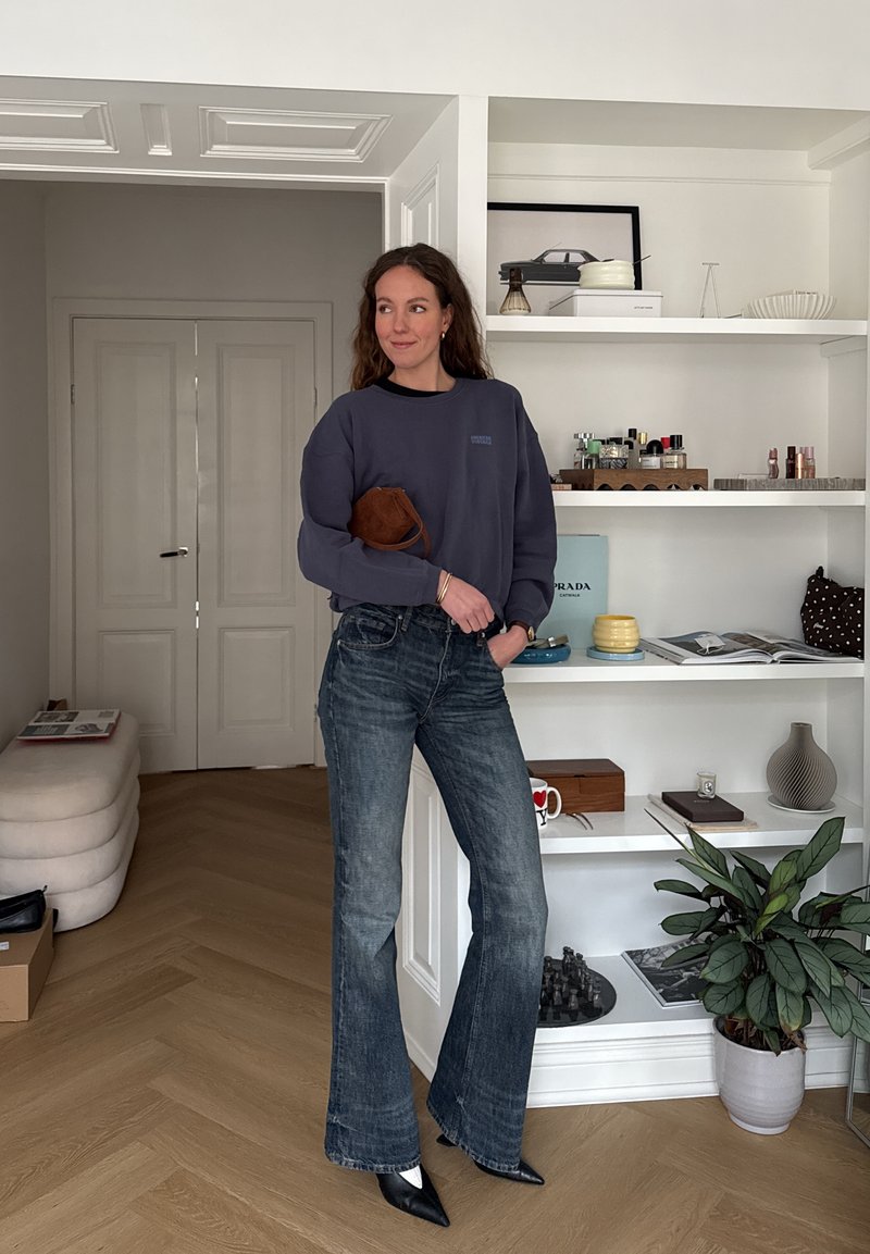 Femme debout à l'intérieur, portant un pull sombre, un jean évasé et des chaussures à bout pointu, tenant un petit sac marron, avec des étagères blanches et des décorations derrière elle.