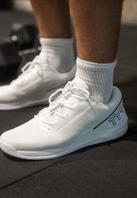 Λευκά αθλητικά παπούτσια με πλέγμα υφής στο πάνω μέρος, διαθέτοντας επίπεδα κορδόνια και μαύρη λογότυπο λεπτομέρεια. Φοριούνται με λευκές κάλτσες με ρίγες.