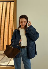 Femme portant une veste en jean surdimensionnée, une chemise blanche, un jean bleu, tenant un sac à main marron, debout à l'intérieur près d'un mur en lattes de bois.