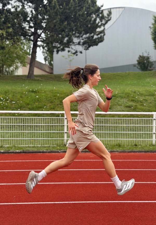Žena u sportskoj odjeći trči na crvenoj vanjskoj stazi s zelenom travom, drvećem i modernom zgradom u pozadini.