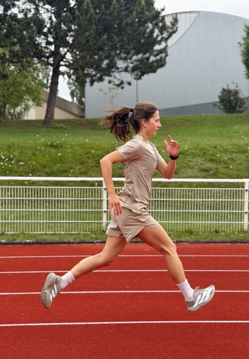 Femme en tenue de sport courant sur une piste extérieure rouge avec de l'herbe verte, des arbres et un bâtiment moderne en arrière-plan.