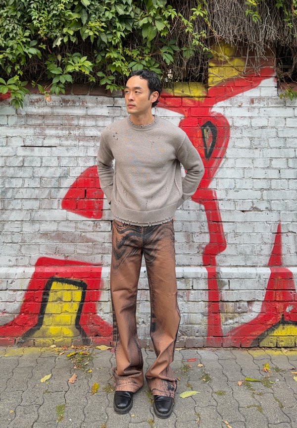 Hombre con suéter beige y pantalones marrones con estampado está apoyado contra una pared de ladrillos blancos cubierta de grafitis, con enredaderas verdes arriba.