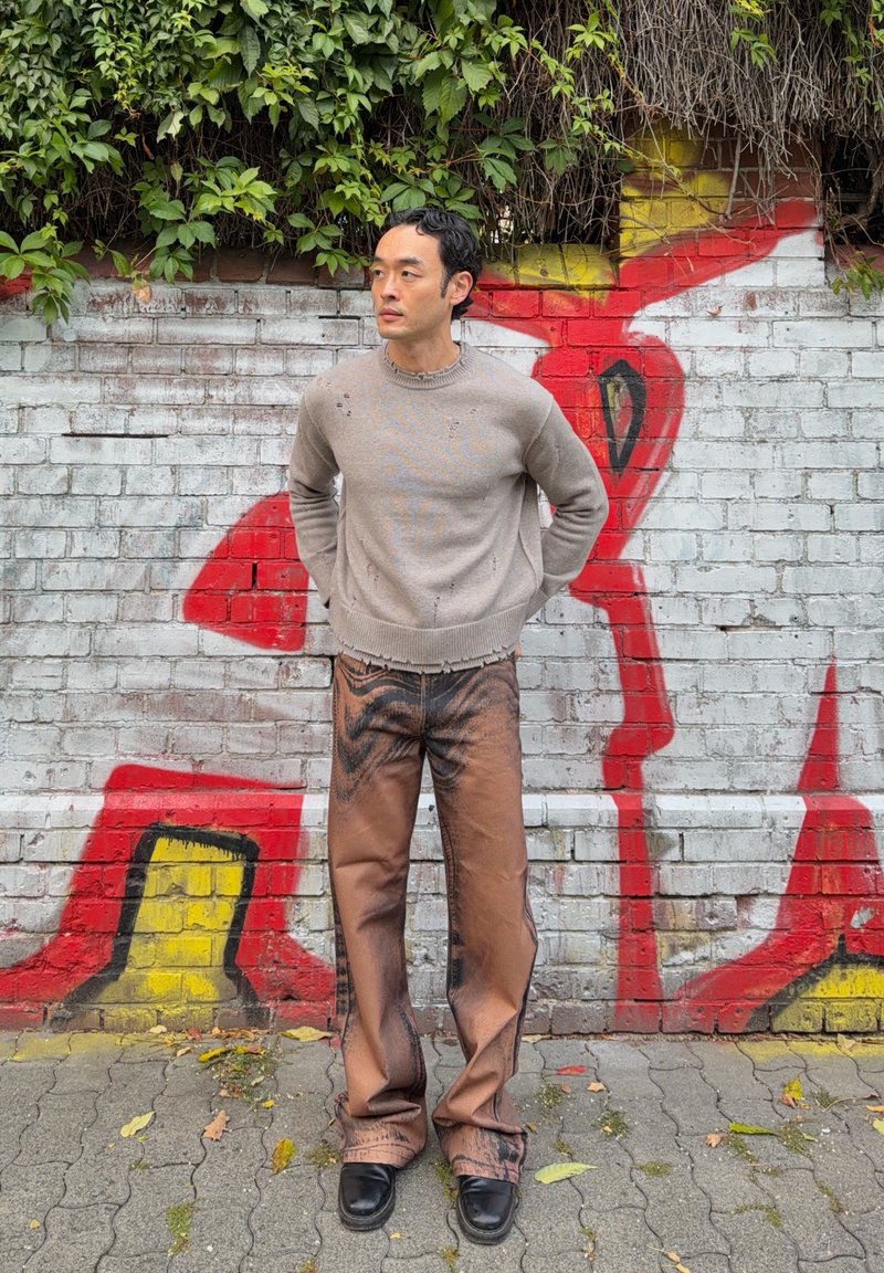 Homme en pull beige et pantalon marron à motifs, debout contre un mur en briques blanches couvert de graffiti avec des vignes vertes au-dessus.