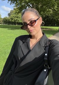 Svart blazer med lange ermer og belte i taljen. Gjennomsiktige solbriller i en mørk nyanse. Svart skulderveske med skinnende detaljer. Grønt gress i bakgrunnen.