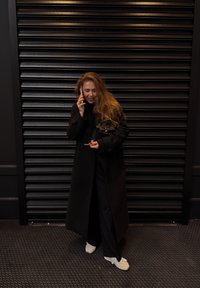 Sort trenchcoat over mørkt outfit, hvide sko, holder en telefon og en pen, stående mod en tekstureret sort metalbaggrund.