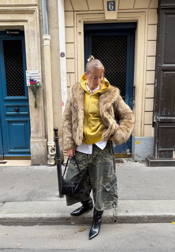 Jachetă din blană artificială, hanorac galben cu fermoar, cămașă albă, pantaloni largi cu model camuflaj, geantă de mână neagră și botine negre cu vârf ascuțit. Ambient urban stradal.