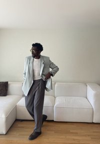 Muž v svetlomodrom saku, bielej košeli, sivých nohaviciach, slnečných okuliaroch a s náušnicou stojí so skríženou jednou nohou v obývačke s bielou pohovkou.