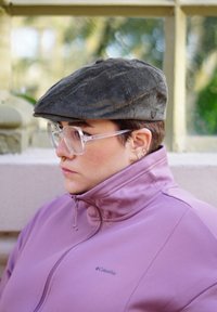 Cappello piatto in corduroy grigio, occhiali rettangolari trasparenti e una giacca zip leggera di colore viola chiaro. L'attenzione è rivolta alla trama e al design del cappello.