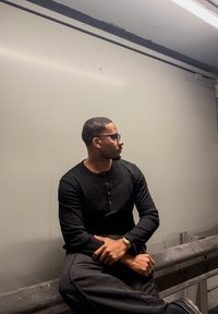 Chemise noire à manches longues côtelée avec un col henley, associée à un pantalon foncé. La personne porte des lunettes et une montre-bracelet en or.