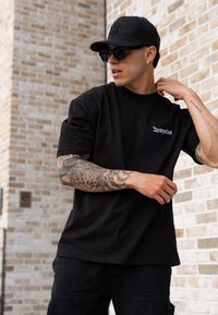 Black cotton t-shirt featuring the "Dropsize" logo. The model is wearing black cargo trousers, sunglasses, and a black cap, showcasing visible tattoos on their arms.