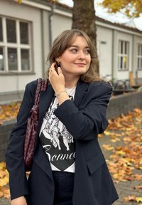 Blazer a righe blu navy sopra una t-shirt con grafica, accessoriate con orecchini a cerchio dorati e una borsa intrecciata color bordeaux, sullo sfondo di fogliame autunnale.