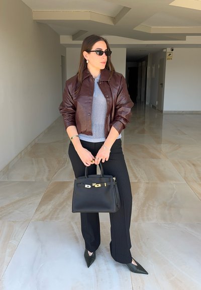 Mujer que lleva chaqueta de cuero marrón, top de punto gris, pantalones negros de rayas, gafas de sol negras, sosteniendo un bolso negro en un pasillo con azulejos.