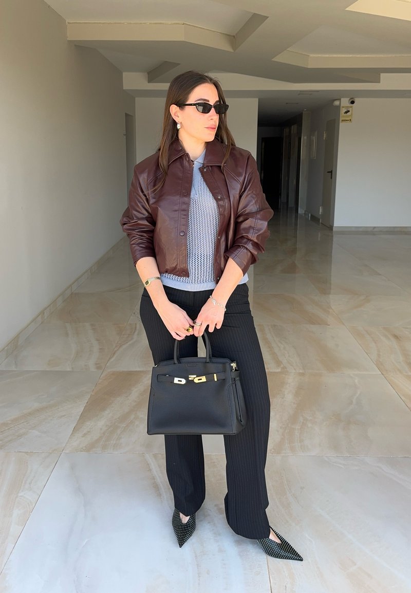 Femme portant une veste en cuir marron, un haut en maille gris, un pantalon noir à fines rayures, des lunettes de soleil noires, tenant un sac à main noir dans un couloir carrelé.