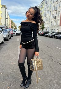 Kobieta pozująca na ulicy w czarnej bluzce bez ramiączek, w szortach z cekinami, w przezroczystych rajstopach, w kozakach za kolano, trzymająca złotą torebkę, w otoczeniu zaparkowanych samochodów i budynków.