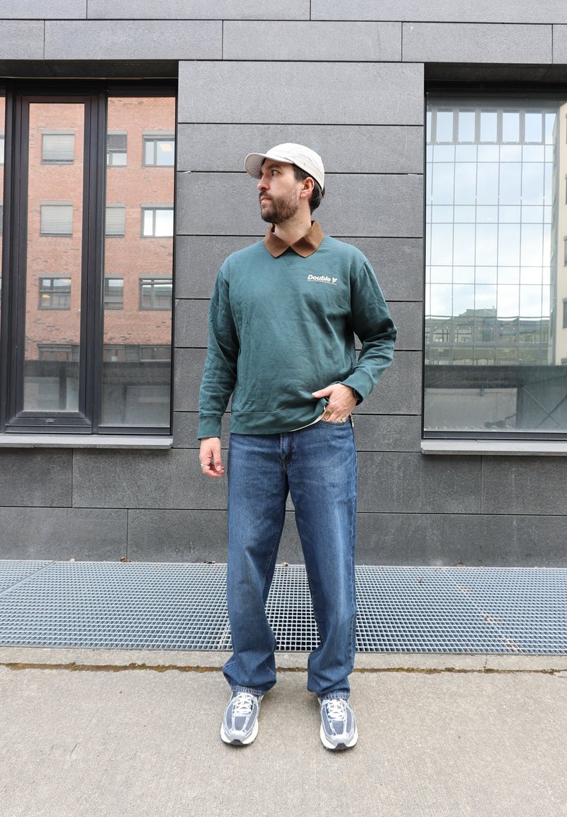 Bărbat purtând șapcă bej, hanorac verde cu logo-ul „Double”, blugi albaștri și adidași, stând în fața unui perete gri placat cu faianță între două ferestre.