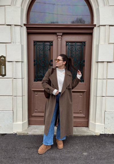 Abrigo de lana marrón, suéter de punto crema, jeans de pierna ancha y zapatos marrones con plataforma. Puerta verde de hierro forjado con un diseño ornamentado.