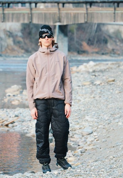 Persona con gorro negro, gafas de sol, chaqueta beige y pantalones acolchados negros de pie en la orilla rocosa de un río cerca del agua, con un puente al fondo.