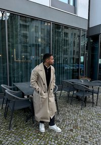 Beige trenchcoat dengan kancing besar, dipakai di atas sweater hitam. Celana hitam dan sepatu sneaker putih. Area makan luar ruangan yang berpaving batu di latar belakang.