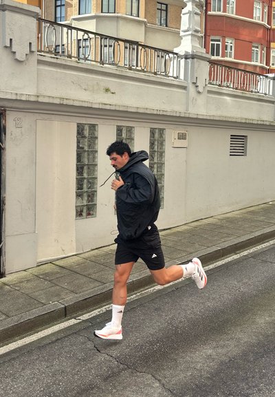 Chaqueta con capucha negra, pantalones cortos negros, zapatillas deportivas blancas con detalles rojos, corriendo por una calle de adoquines con edificios de fondo.