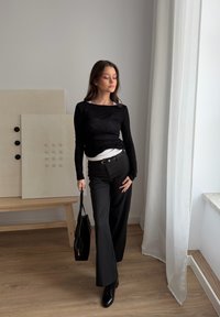 Jeune femme en haut et pantalon noirs tenant un sac noir, debout près d'un rideau blanc et d'un banc en bois avec des panneaux texturés.