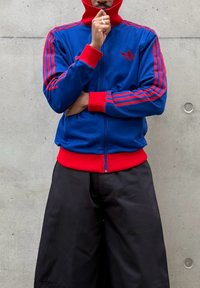 Άτομο με μπλε και κόκκινο μπουφάν Adidas με κουκούλα, το χέρι ακουμπάει το πρόσωπο, φορά φαρδιά μαύρη παντελόνια, στέκεται μπροστά σε τσιμεντένιο τοίχο.