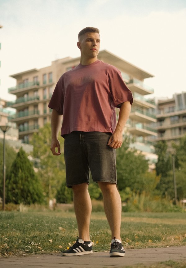 Jeune homme en chemise bordeaux ample, short noir et baskets, debout sur un sentier dans un parc avec des immeubles d’appartements modernes en arrière-plan.