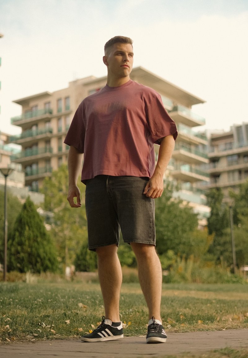 Jeune homme en chemise bordeaux ample, short noir et baskets, debout sur un sentier dans un parc avec des immeubles d’appartements modernes en arrière-plan.