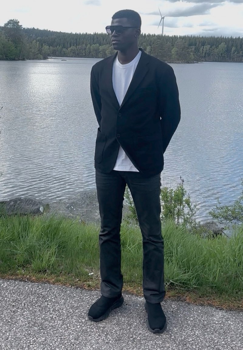 Homme portant un blazer noir, une chemise blanche, des lunettes de soleil et un pantalon noir debout sur un chemin au bord d’un lac avec des arbres et une éolienne en arrière-plan.