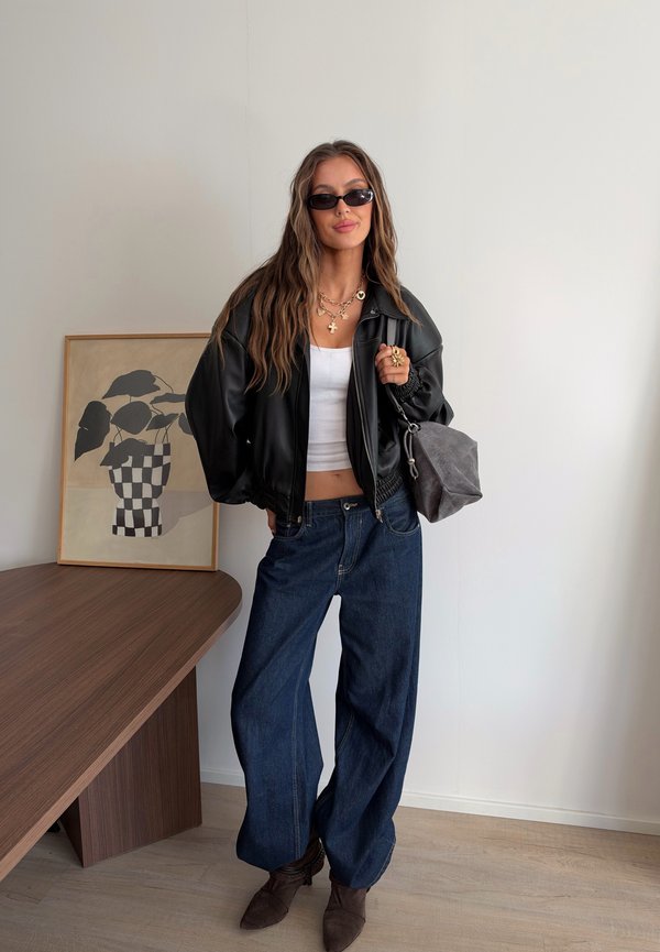 Femme portant une veste en cuir noire, un crop top blanc, un jean large bleu, des bottes marron, des lunettes de soleil, des bijoux en or, et tenant un sac à main gris à l'intérieur.