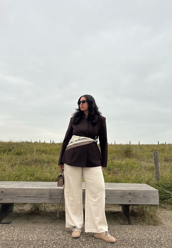 Brun blazer med satinbælte, cremefarvede brede bukser og brune sko. Accessoriseret med mørke solbriller og en lille håndtaske. Udendørs omgivelser.
