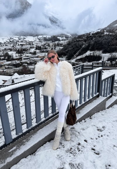 Mujer con abrigo de piel blanco, pantalones blancos y botas, de pie en un balcón nevado con vistas a un pueblo cubierto de nieve y montañas envueltas en niebla.