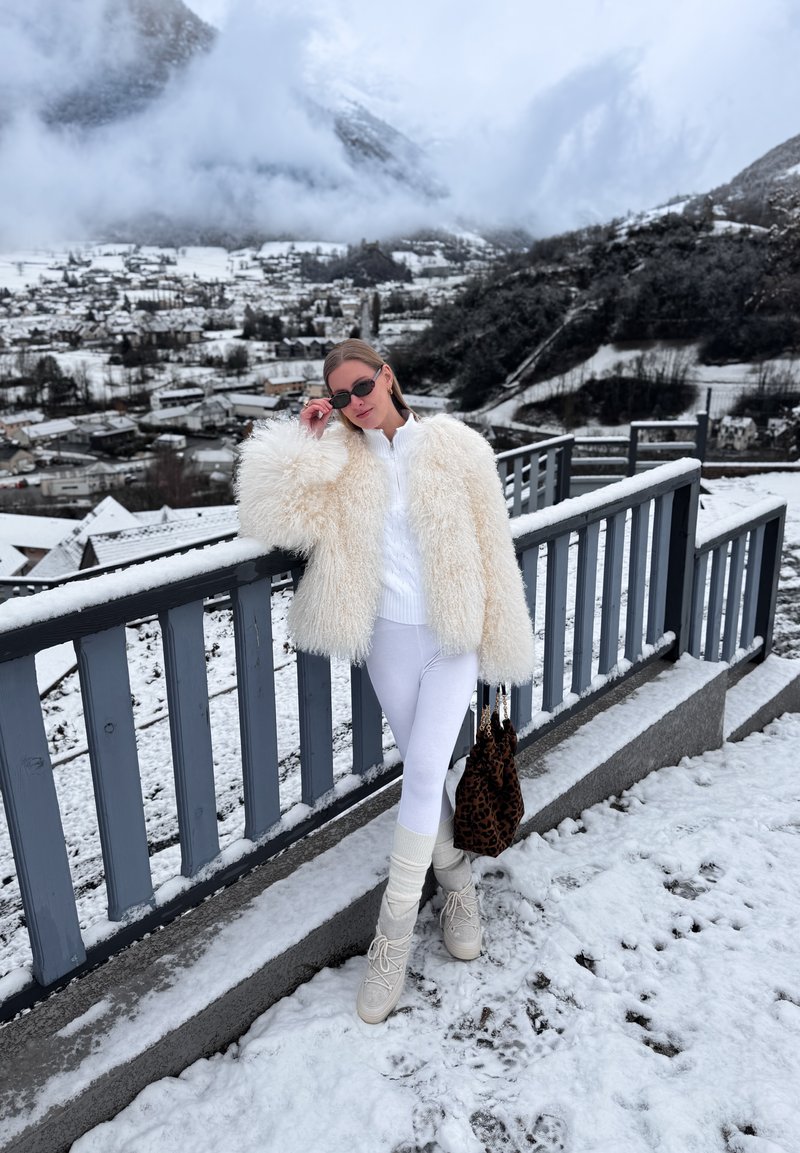 Ženska v belem krznenem plašču, belih hlačah in škornjih stoji na zasneženem balkonu, ki gleda na zasneženo vas in gore v megli.