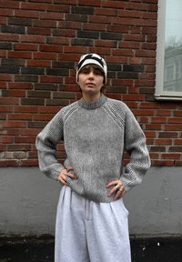Suéter de punto grueso en rayas blancas y negras, con escote redondo y mangas raglán. Combinado con pantalones deportivos grises holgados y un gorro estampado.