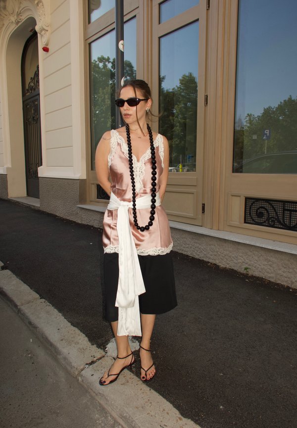 Femme portant un caraco en satin rose avec bordure en dentelle, des culottes noires, une ceinture blanche, un long collier noir perlé, des lunettes de soleil et des sandales se tient sur le trottoir.