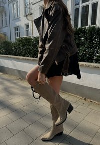 Rjava usnjena jakna z detajlnim šivanjem, v kombinaciji z črno obleko in svetlobezžnimi gamšastimi čizmami do kolen z debelim peto.