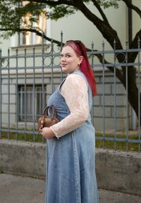 Farmerruha testhez simuló felsőrésszel, bővülő szegéllyel és csipkés hosszú ujjakkal. Egy kis barna kézitáskát tart. A háttérben szürke kerítés és zöld fal látható.