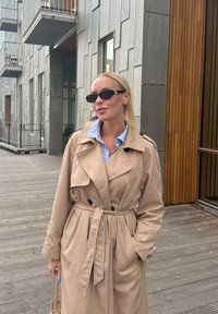 Bež trench coat, mille vööga, kahekordsete nööpidega ja laia kraega; koos tumedate päikeseprillide ja helesinise särgiga.