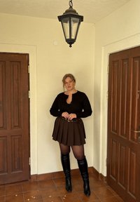 Camisa negra de manga larga, falda marrón con pliegues y cinturón, medias transparentes, botas negras hasta la rodilla; posando contra una pared de color claro.