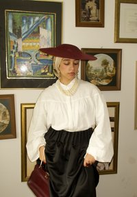 Czerwona szerokokrawędziowa kapelusz, biała bluzka z bufkami, czarna satynowa spódnica, naszyjnik z pereł oraz bordo torebka na tle oprawionych dzie�ł sztuki.