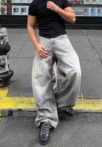 Széles szárú, világosszürke farmernadrág elhalványult textúrával és festékfröccsenésekkel. Fekete, fűzős bakancsokkal és szűk fekete inggel párosítva.