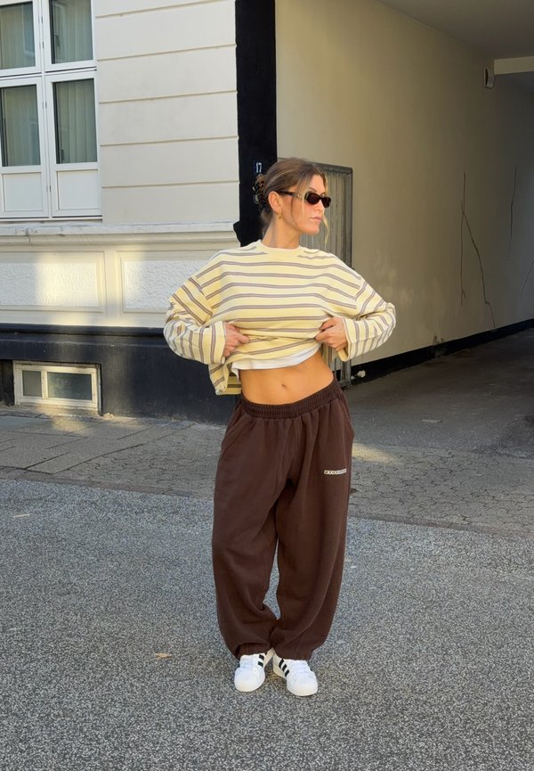 Mujer con gafas de sol levanta un jersey a rayas para mostrar el abdomen, de pie en una calle cerca de la entrada de un edificio beige.