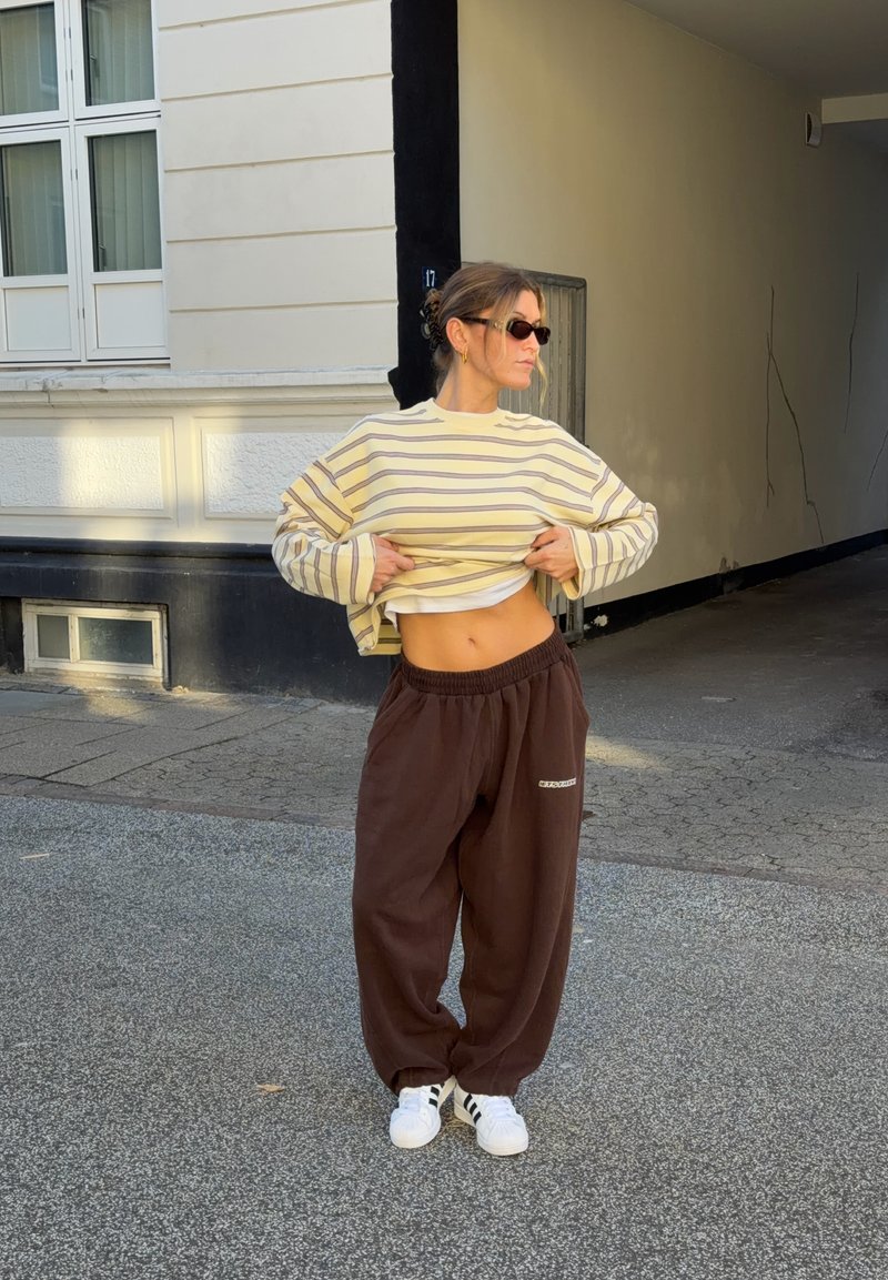 Femme portant des lunettes de soleil soulève un pull rayé pour montrer le ventre, debout dans une rue près de l'entrée d'un bâtiment beige.
