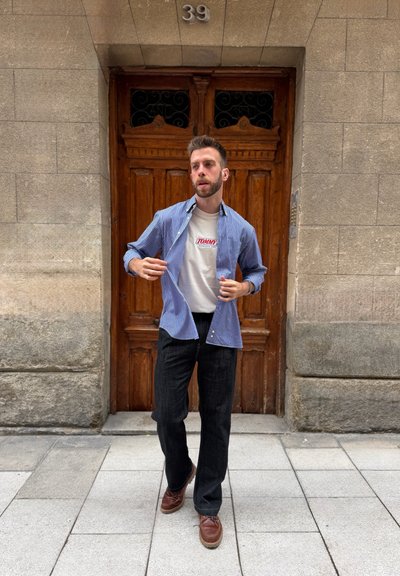 Hombre de pie frente a una puerta de madera con el número 39, vistiendo una camisa de rayas azules sobre una camiseta blanca Tommy, pantalones negros y zapatos marrones.