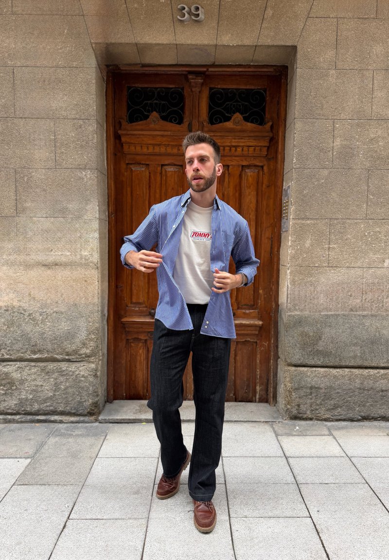 Mann steht vor einer Holztür mit der Nummer 39, trägt ein blau gestreiftes Hemd über einem weißen Tommy-T-Shirt, schwarze Hose und braune Schuhe.