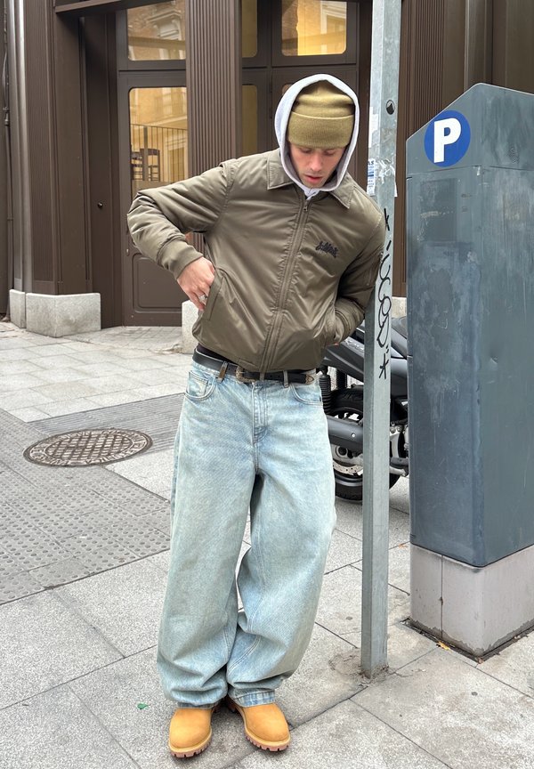 Jeune homme portant une veste marron, un bonnet beige, un jean large clair et des bottes jaunes, debout à côté d'un parcmètre sur un trottoir urbain.
